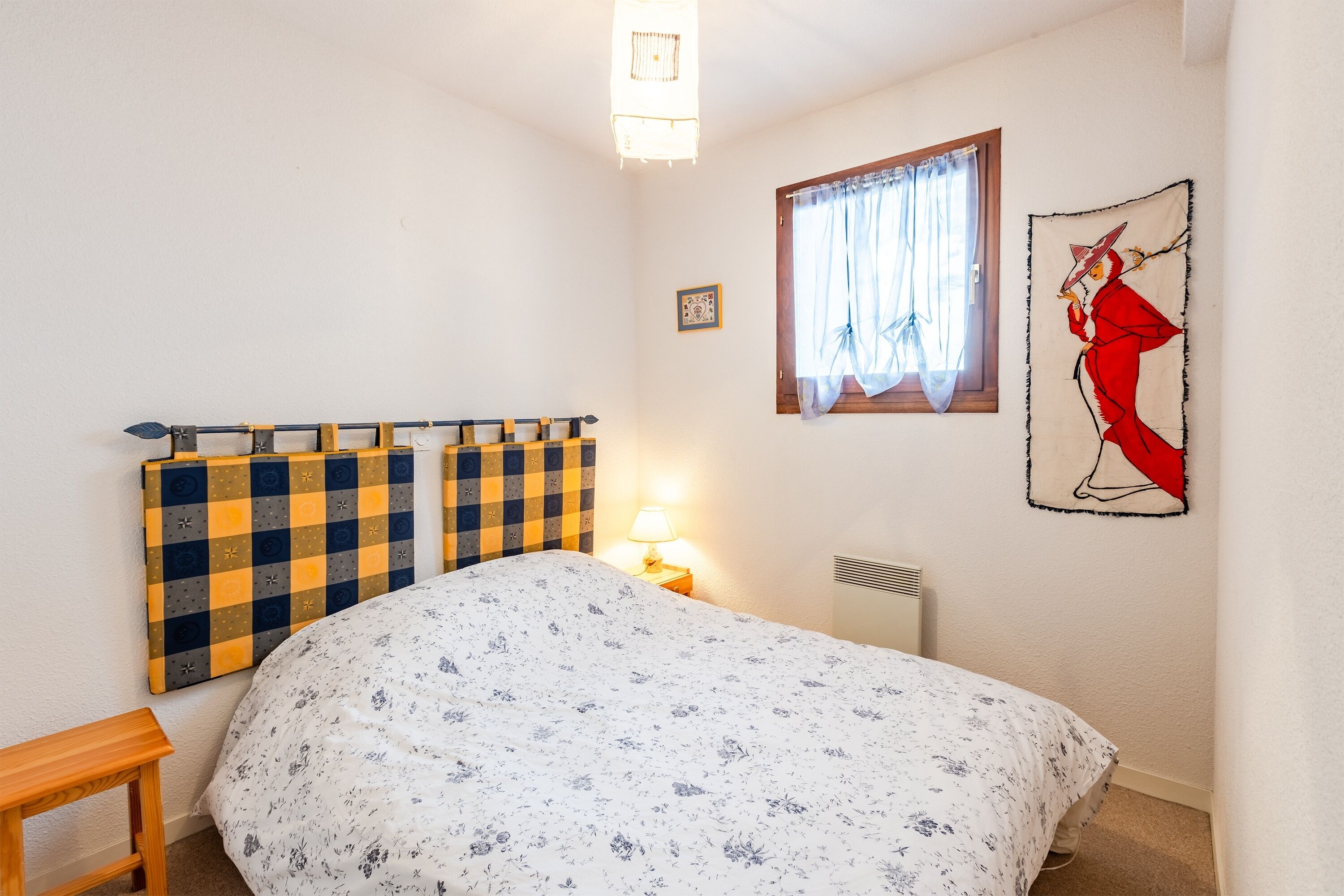 Una camera da letto con un letto, una testiera a quadri, una finestra con tende, una lampada a parete e un quadro incorniciato.