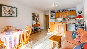 Una sala de estar acogedora con un sofá, una mesa de comedor con sillas, una zona de cocina con armarios de madera y electrodomésticos, y una estantería.