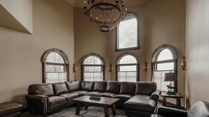 Ein geräumiges Wohnzimmer mit einer großen Eckcouch, einem Couchtisch und einer Deckenleuchte. Drei Rundbogenfenster sorgen für natürliches Licht.