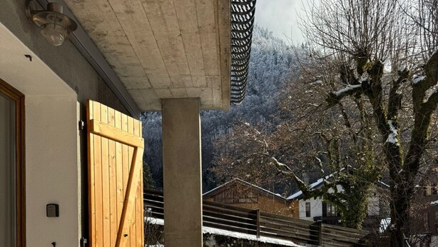 A wooden deck leading to a building with a large wooden door, a covered outdoor area, and a snowy landscape in the background.