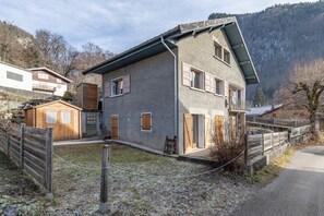 A two-story house with a sloped roof, wooden shutters, and a small wooden shed in the yard. - Samoëns – Apartment for 6 with Terrace, Garden & Panoramic Views (Samoëns)
