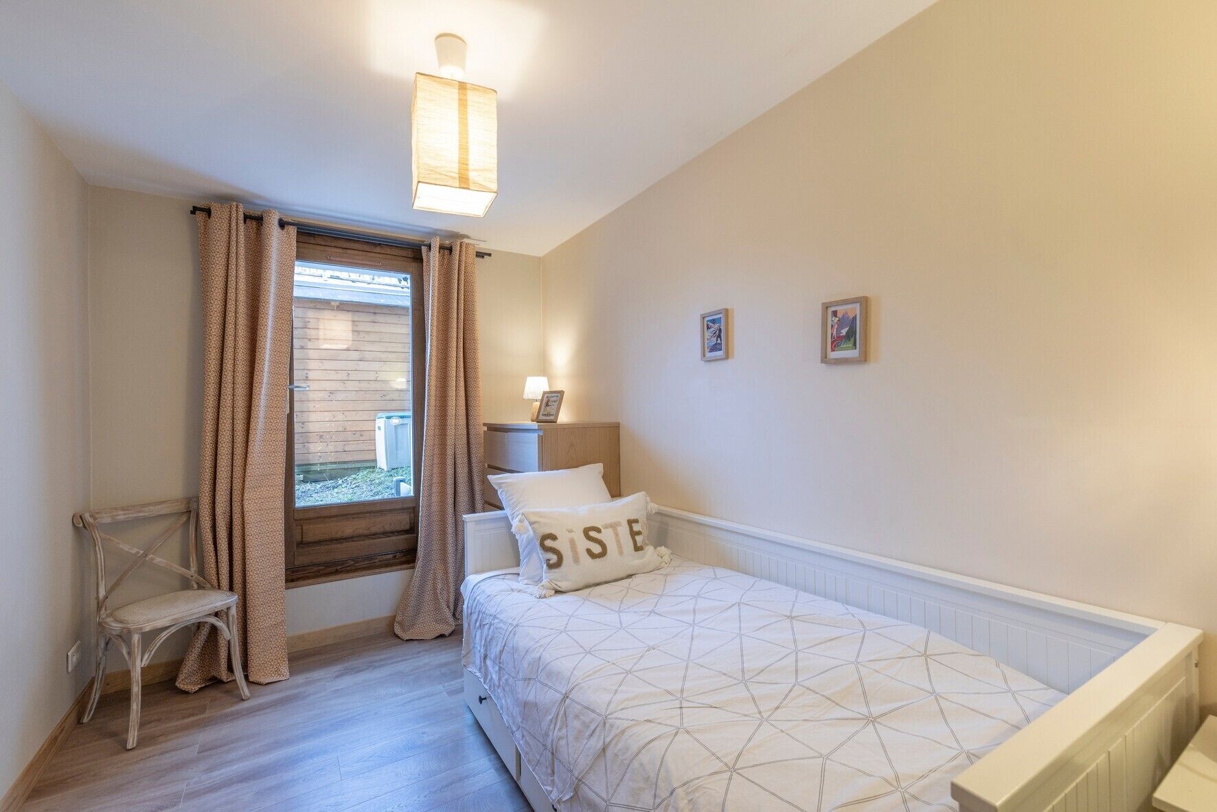 Ein Schlafzimmer mit Bett, Sessel, einem Fenster mit Vorhängen, einem Nachttisch mit Lampe und zwei gerahmten Bildern an der Wand.