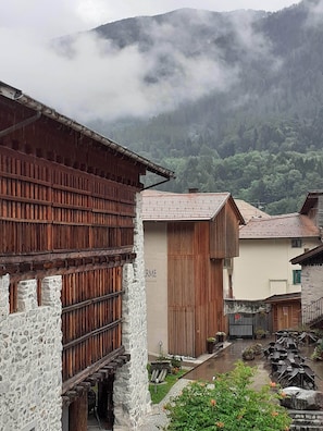 Um edifício rústico com fachada de madeira e paredes de pedra, um pequeno jardim com flores e montanhas ao fundo.