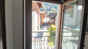 Blick durch eine Glastür auf ein Gebäude mit Balkon, eine Steinmauer und einen Parkplatz mit Autos.