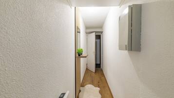 Un couloir étroit aux murs blancs, avec un plancher en bois et un tapis en fourrure blanche. Une petite plante est posée sur une étagère, et un thermostat est fixé au mur.