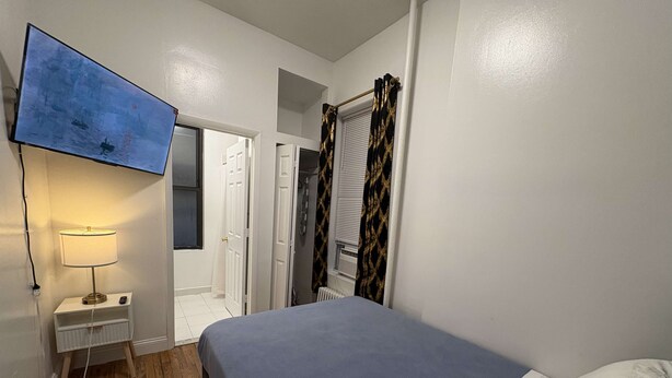 Un dormitorio con cama, televisor montado en la pared, mesita de noche con lámpara y puerta que da a un baño.