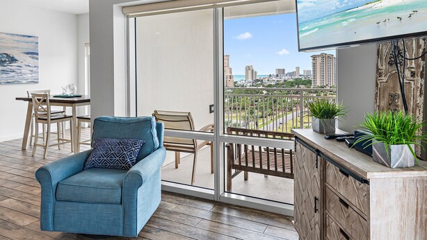 A balcony with a city view, a sofa, a dining table with chairs, and a TV mounted on the wall. - 🏖️Beautiful Views-Lagoon Pool-Hot Tub-Walk2Beach-Luau II- By the Sea☀️ (Miramar Beach)