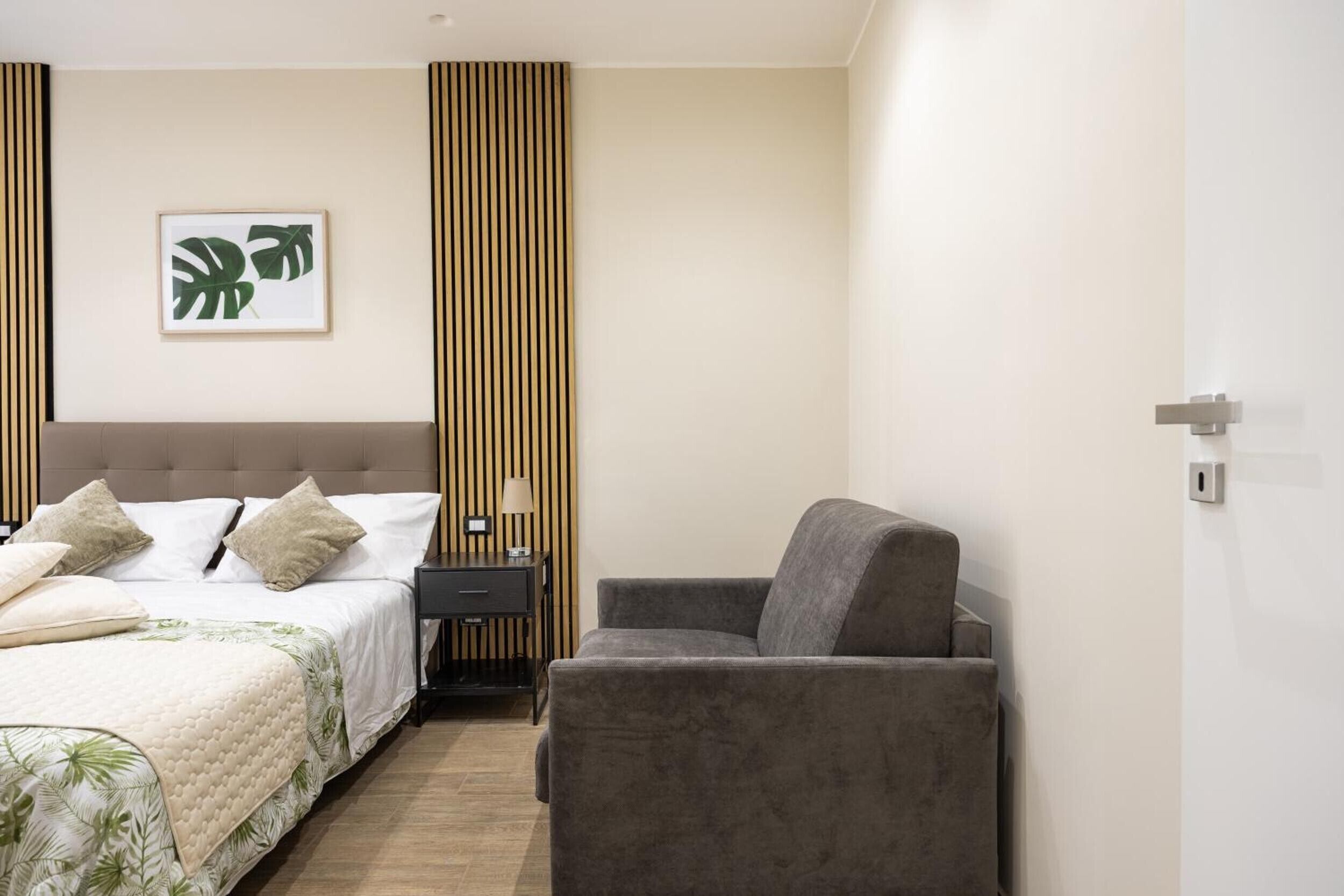 A hotel room with a bed, a grey armchair, a nightstand, and a framed picture on the wall.