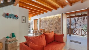 Een kamer met een houten plafond, een stenen muur, een rode bank, rieten manden en een zithoek met stoelen en een tafel.