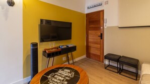 Une pièce avec un mur jaune, un téléviseur fixé au mur, une table en bois avec un plateau noir, deux tabourets noirs, une porte en bois et une petite plante.