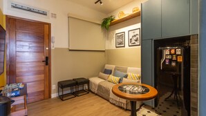 Un espace de vie compact doté d’une porte en bois, d’un canapé avec des coussins, d’une petite table ronde, d’un réfrigérateur et d’une étagère murale agrémentée d’objets décoratifs.