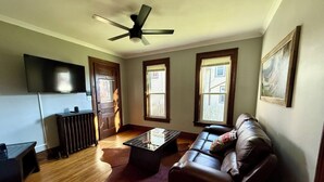 A living room with a brown leather sofa, a glass coffee table, a wooden floor, a ceiling fan, and a flat-screen TV mounted on the wall. - Western-Themed & Conveniently-Located Apartment in Tonawanda (Tonawanda)