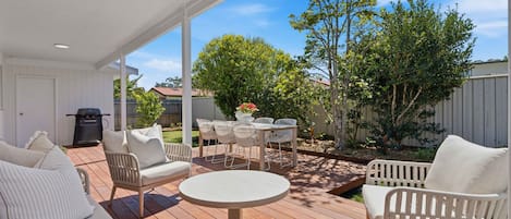 Un patio techado al aire libre con una mesa redonda y sillas, una parrilla y una cerca blanca.