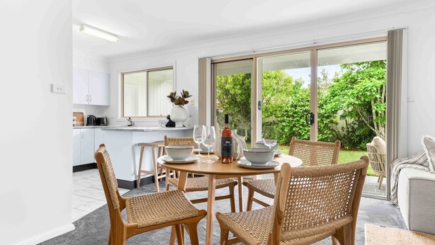 Une cuisine moderne avec des meubles blancs, un coin repas doté de chaises en osier et une grande porte-fenêtre coulissante donnant sur un jardin.