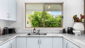 Une cuisine moderne avec des meubles blancs, un évier à deux bacs, un grille-pain, une machine à café et une fenêtre donnant sur un paysage verdoyant.
