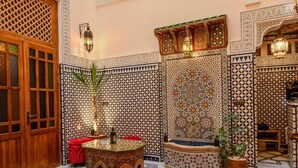 Une pièce aux motifs complexes sur les carreaux, une porte en bois, une petite table avec une lampe et un pouf rouge.