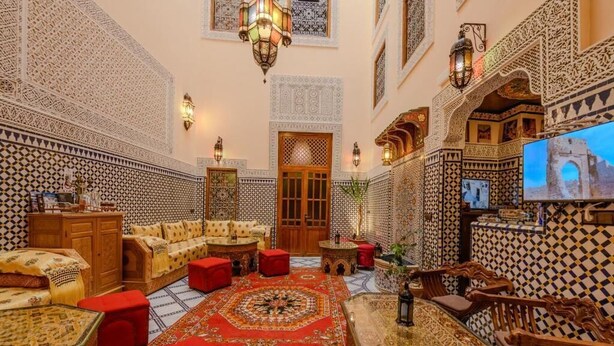 Un intérieur marocain traditionnel avec un espace salon central, des murs richement décorés et un écran de télévision.
