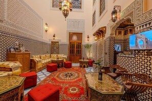 Un interior tradicional marroquí con una zona de estar central, paredes ornamentadas y una pantalla de televisión.