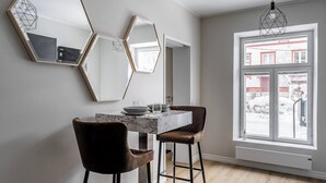 Ein Essbereich mit einem Marmortisch und zwei Stühlen, einem geometrischen Spiegel an der Wand und einem großen Fenster mit Blick auf ein Gebäude.