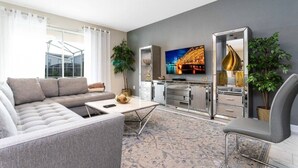 Un salon moderne avec un canapé gris, une table basse, un téléviseur fixé au mur et un coin cuisine doté de placards et d’une plante.