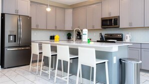 Une cuisine moderne avec des meubles gris, un réfrigérateur en acier inoxydable, un four à micro-ondes et un coin bar avec des chaises blanches.