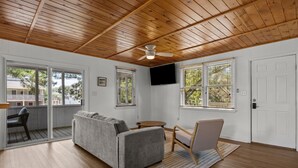 A room with a wooden ceiling, a gray sofa, a wooden chair, a small round table, and a TV mounted on the wall. - Easy Walk to Beach & Nags Head Shops, Restaurants (Nags Head)