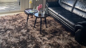 Ein Wohnzimmer mit einem schwarzen Ledersofa, einem kleinen runden Tisch mit Blumen in einer Vase, einer Glastür, die auf einen Balkon führt, und einem Bild an der Wand.