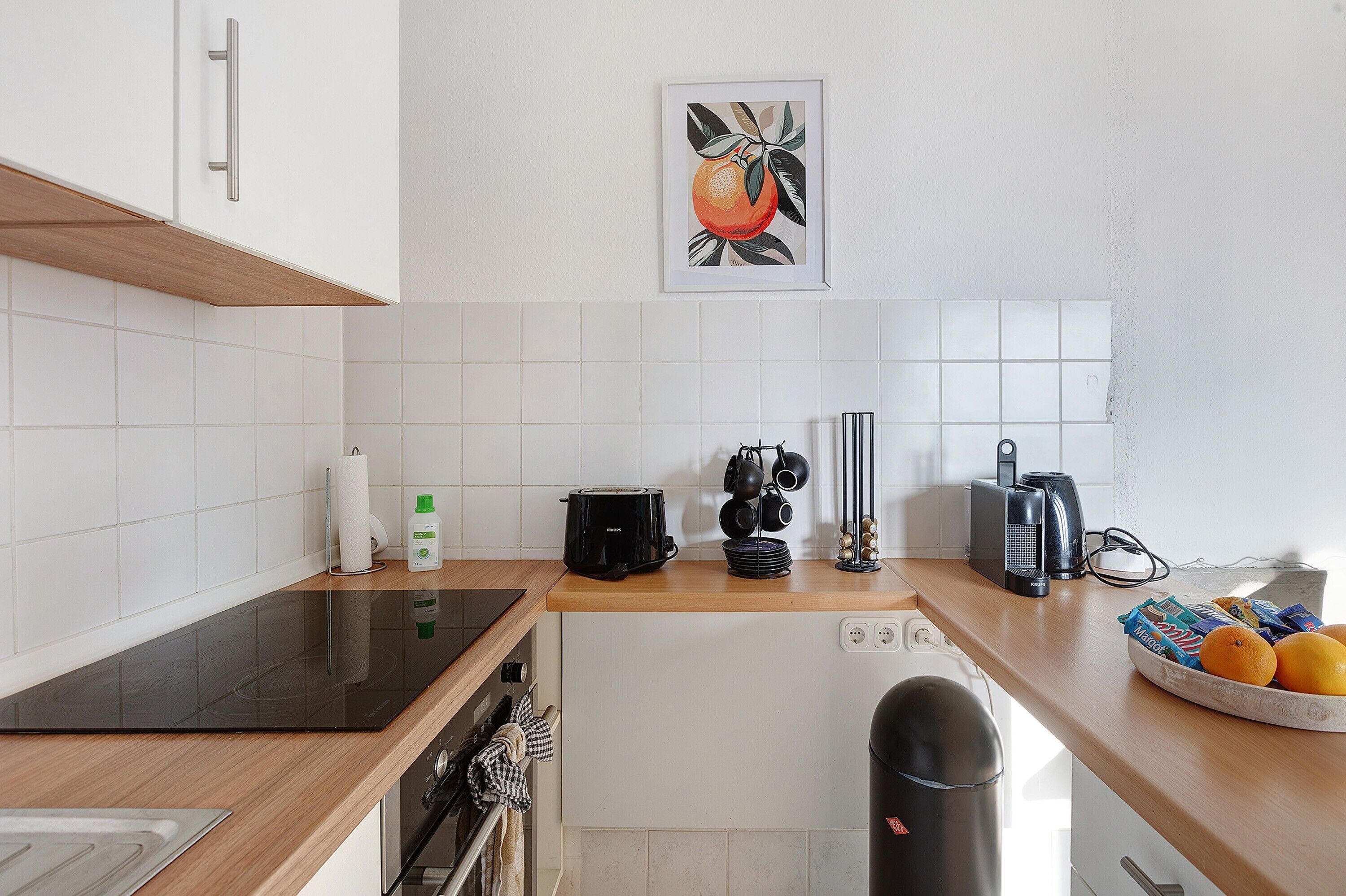 A modern kitchen with white tiles, a black induction cooktop, a coffee maker, and a kettle.