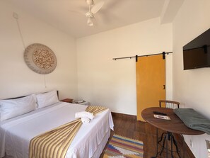 A hotel room with a bed, a small table with a chair, a TV mounted on the wall, and a door leading to another room. - Pousada Garden House Tiradentes (Tiradentes)