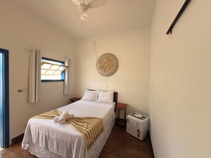 A bedroom with a bed, a ceiling fan, a window with curtains, a wall-mounted decorative plate, and a small heater. - Pousada Garden House Tiradentes (Tiradentes)