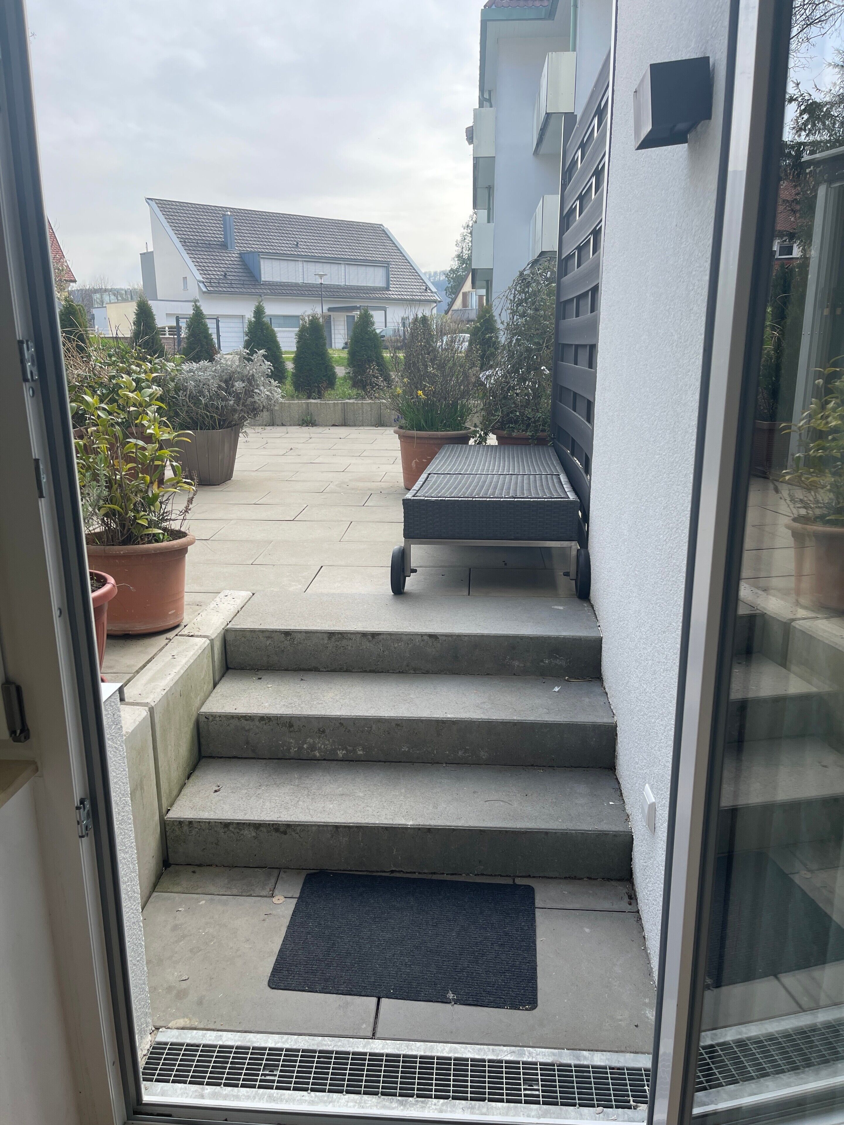 A view through a glass door to a set of concrete steps leading to a small outdoor area with a bench and potted plants.