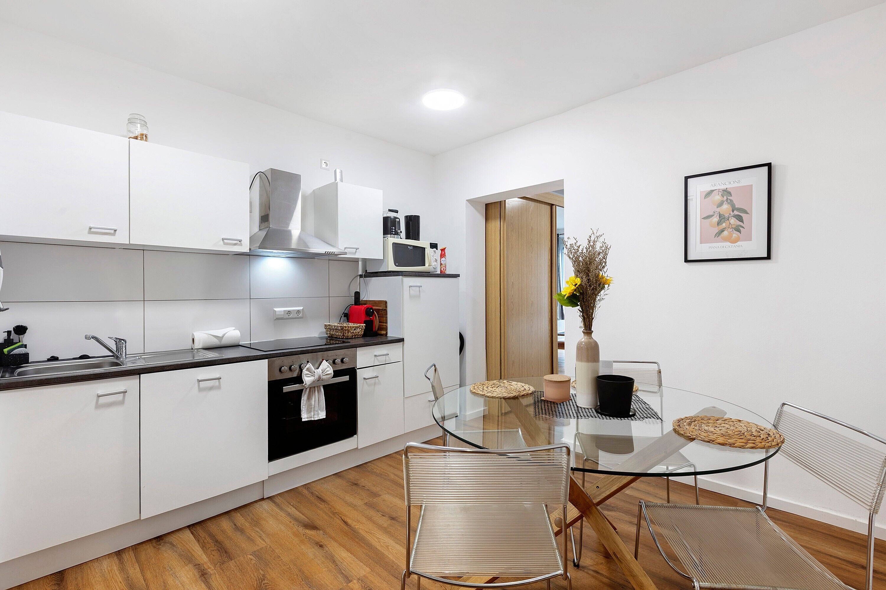 A modern kitchen with white cabinets, a microwave, and a stove. There is a dining area with a glass table and metal chairs.