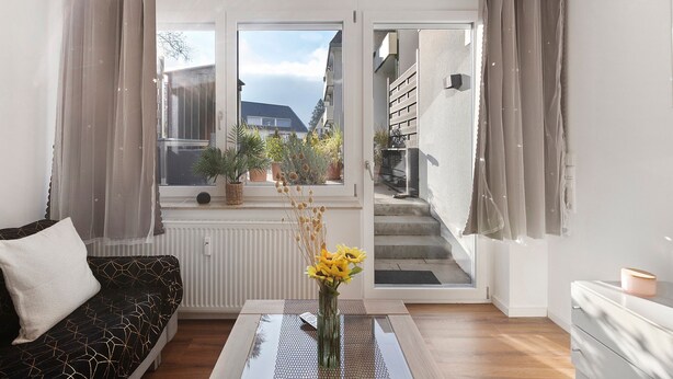 Un salon moderne avec un canapé, une table basse ornée d’un vase de fleurs, une vue sur un balcon accessible par une porte coulissante, et un escalier menant à l’extérieur.