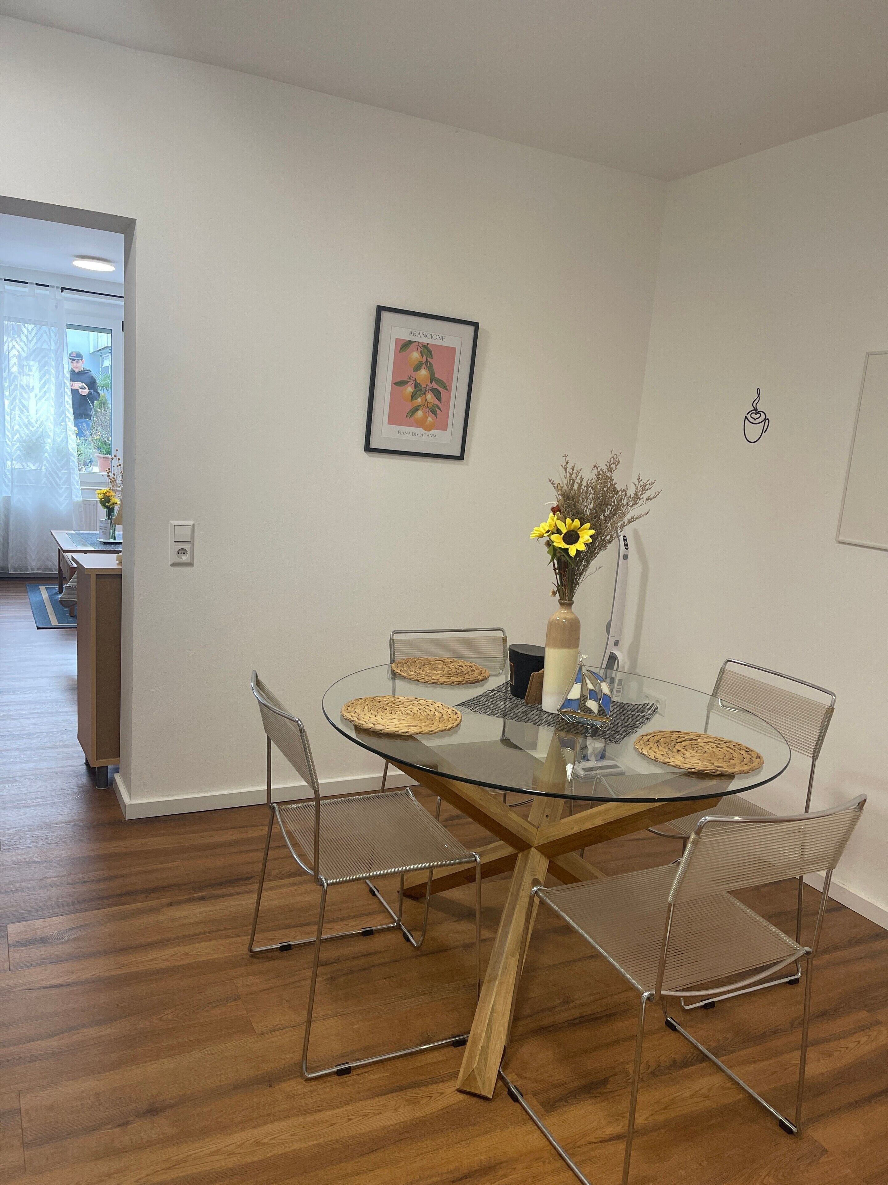 Un coin repas avec une table en verre, des chaises en métal, un vase de fleurs et un tableau encadré au mur.