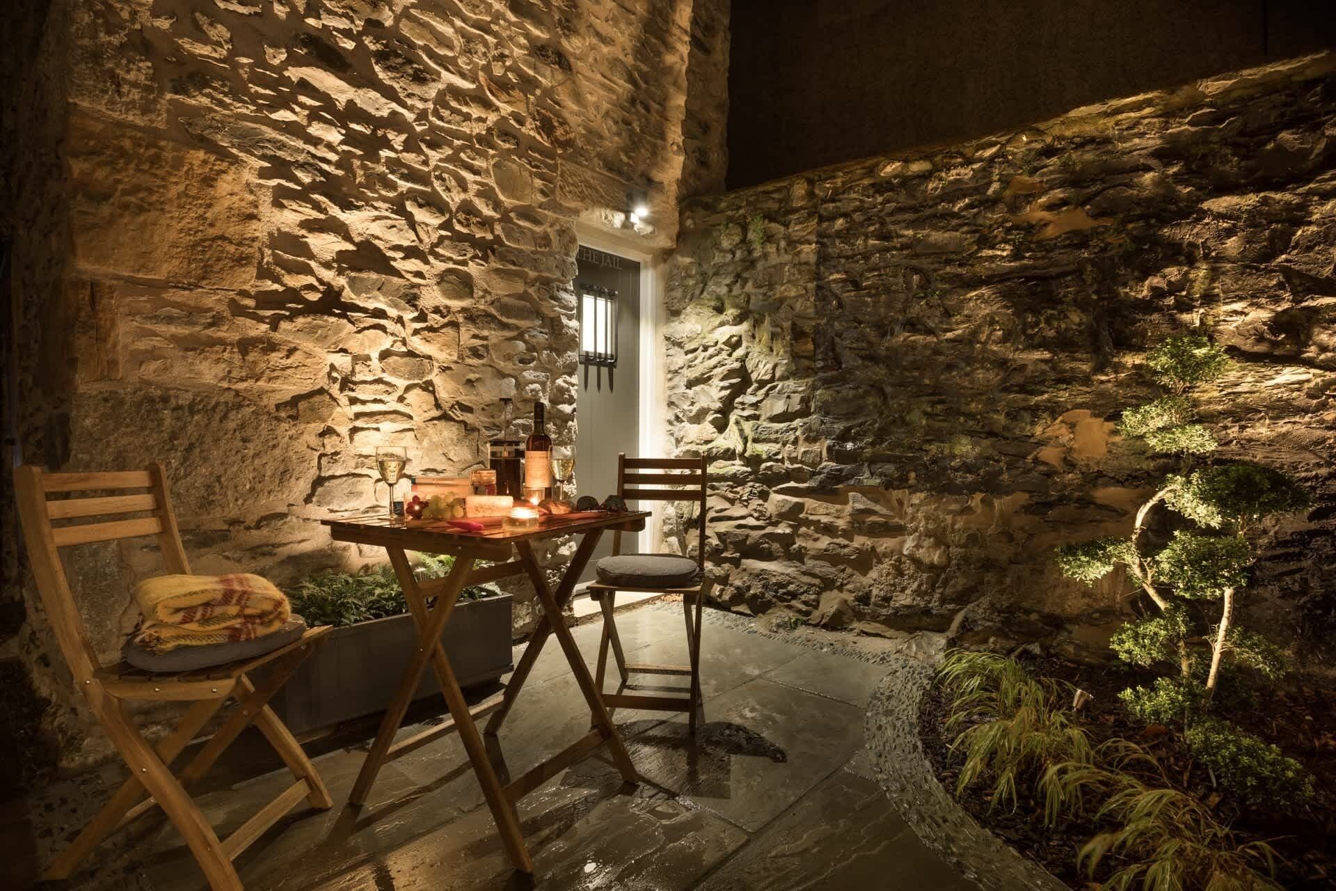 Un espace extérieur aménagé pour les repas, avec des murs en pierre, une table dressée pour deux personnes, une bouteille de vin et une bougie.