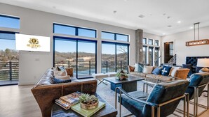 A spacious living room with a large window overlooking a scenic view, a comfortable seating area with a sofa and chairs, and a coffee table with magazines and a plant. - Lakeview Penthouse Loft w/ Pool, Hot Tub, Game (Osage Beach)