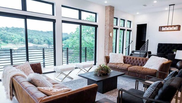 A spacious living room with leather sofas, a coffee table, and large windows offering a view of a forested area. - Lakeview Penthouse Loft w/ Pool, Hot Tub, Game (Osage Beach)