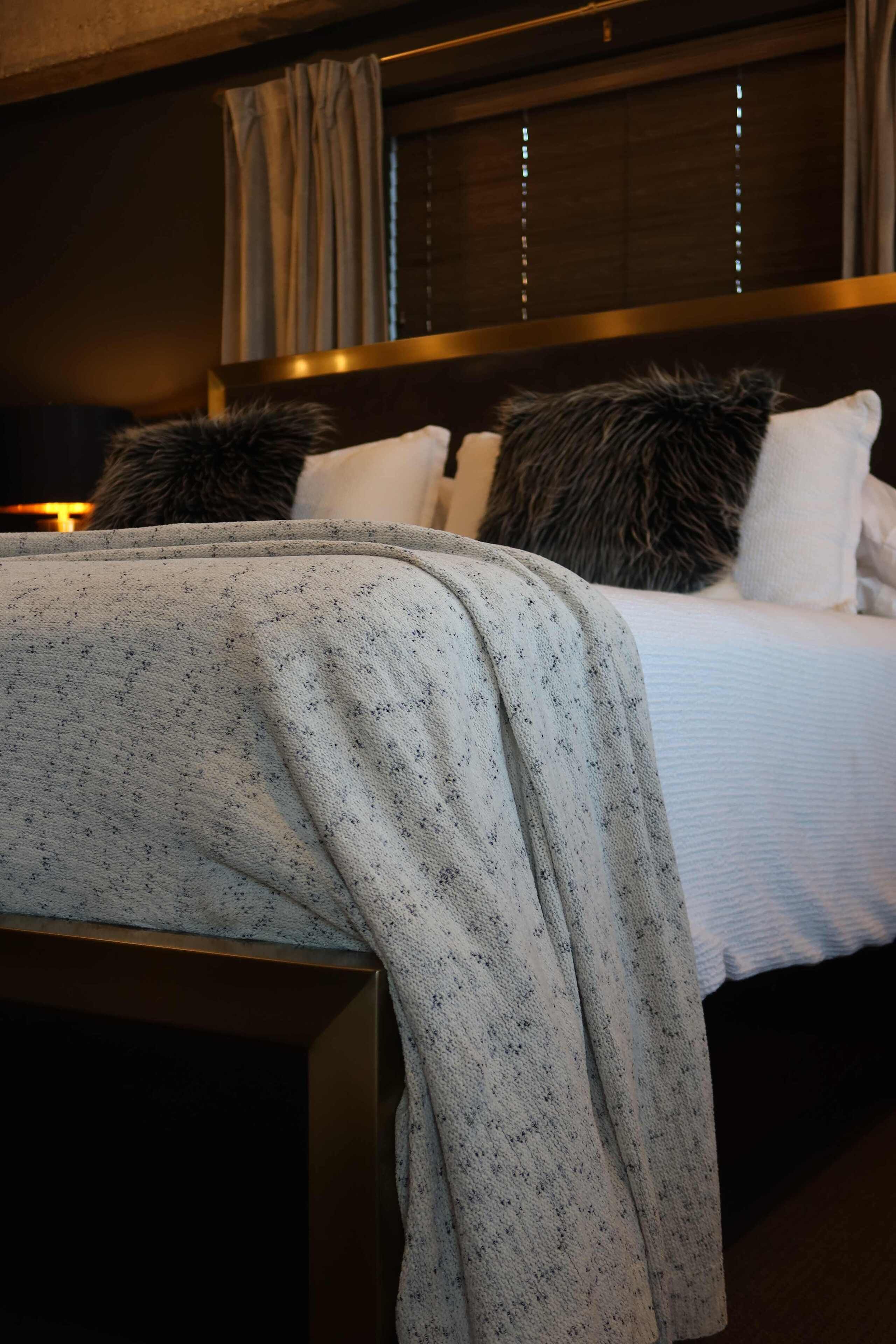 A neatly made bed with a white comforter and a grey blanket, a wooden headboard, and a window with curtains.