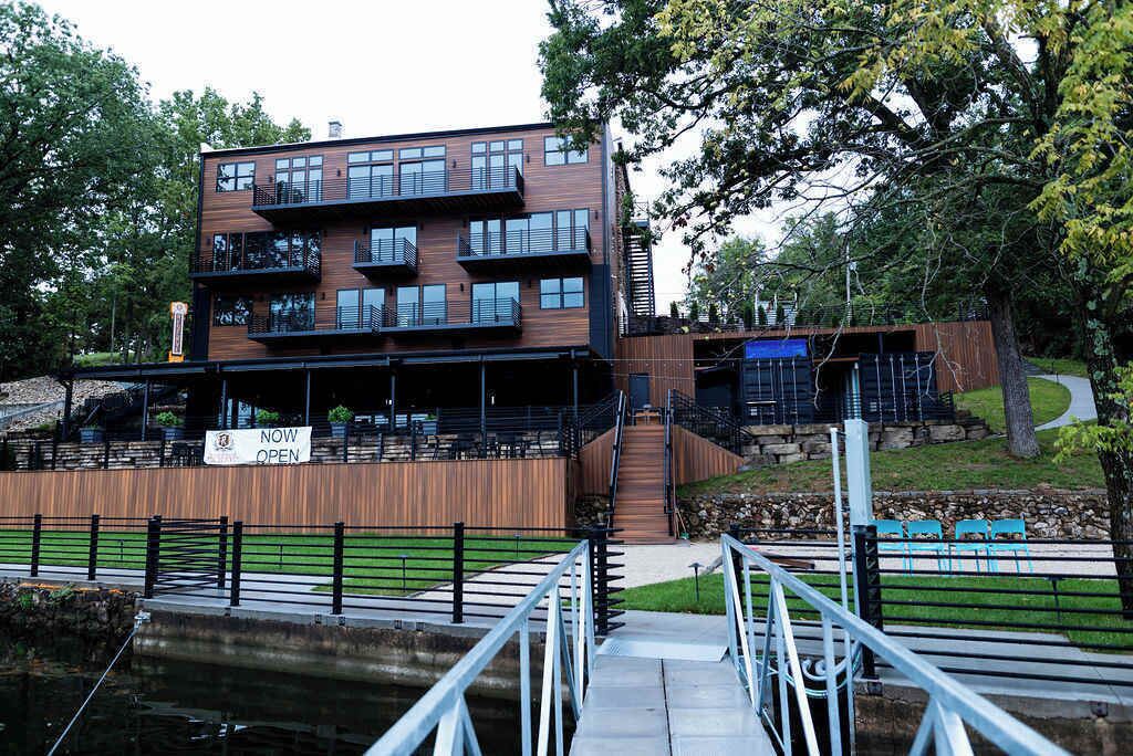A modern multi-story building with balconies, a sign indicating it is now open, and a wooden deck area with chairs.