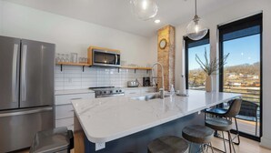 A modern kitchen with a large island, stainless steel appliances, and a view of a river and buildings. - Lakeview Loft: Stylish Retreat with Hot Tub & Pool (Osage Beach)