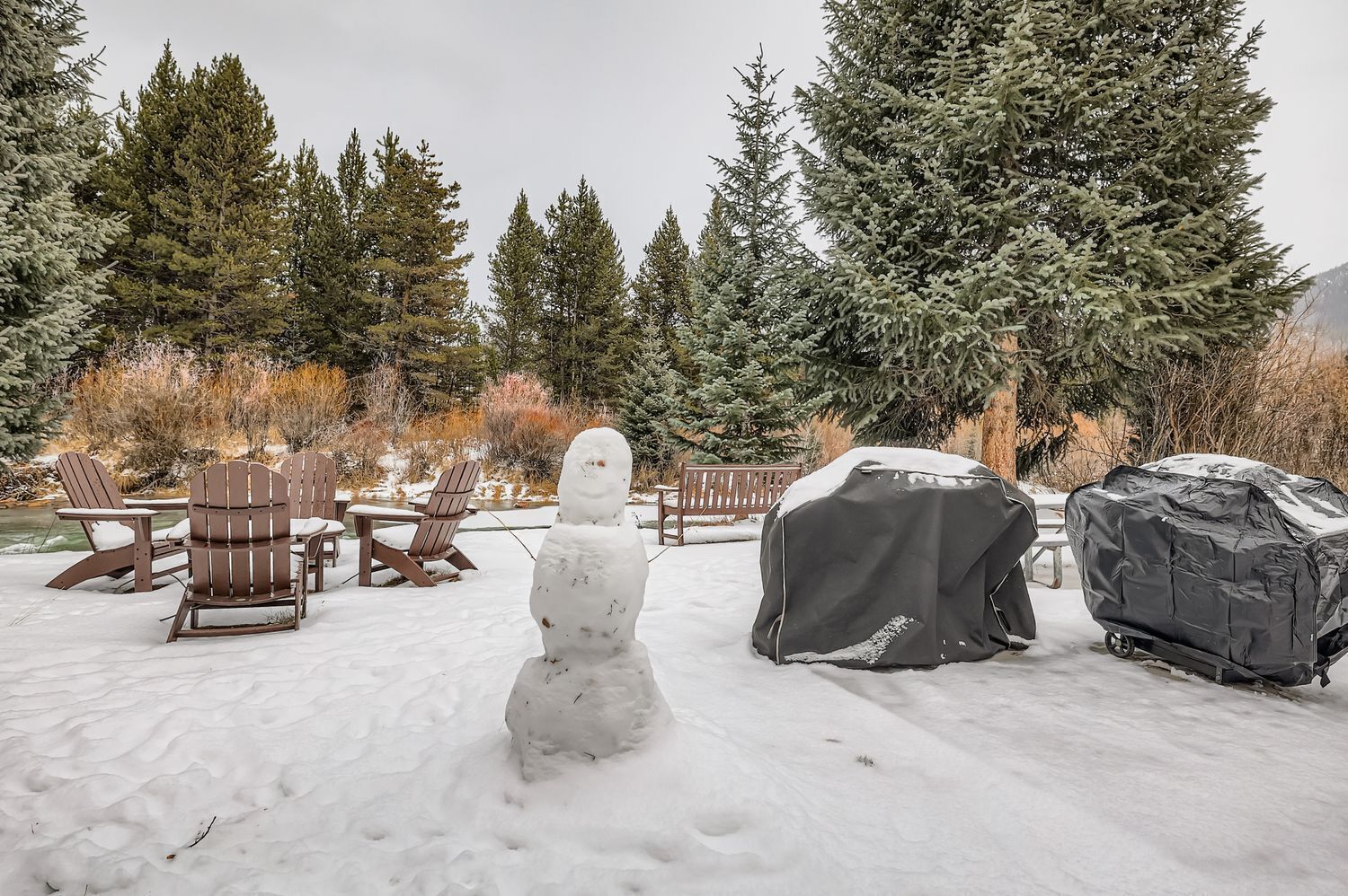 Un muñeco de nieve en un espacio exterior nevado con sillas Adirondack y una parrilla cubierta.