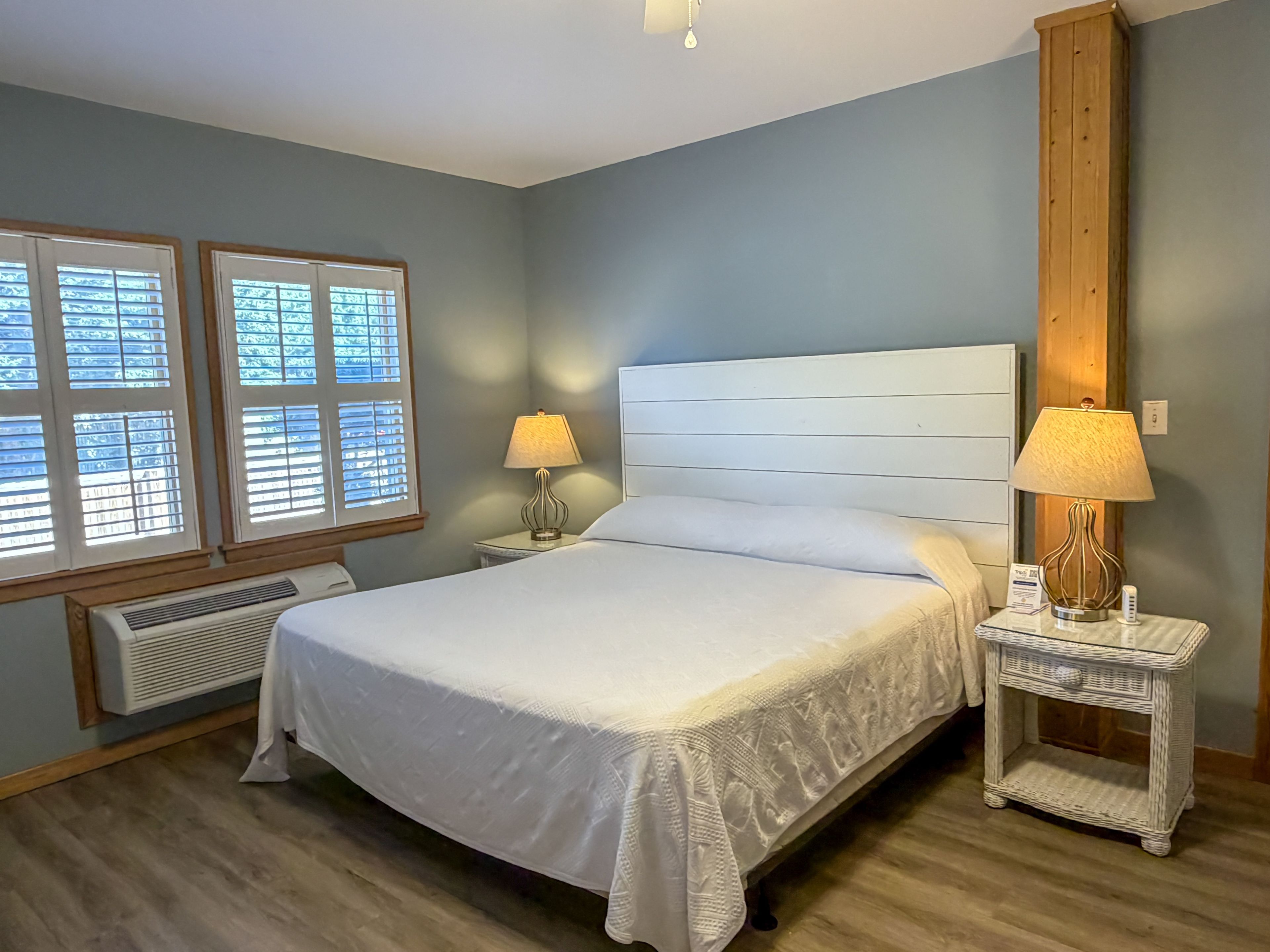 A bedroom with a large bed, two bedside tables with lamps, a window with blinds, and a wall-mounted heater.