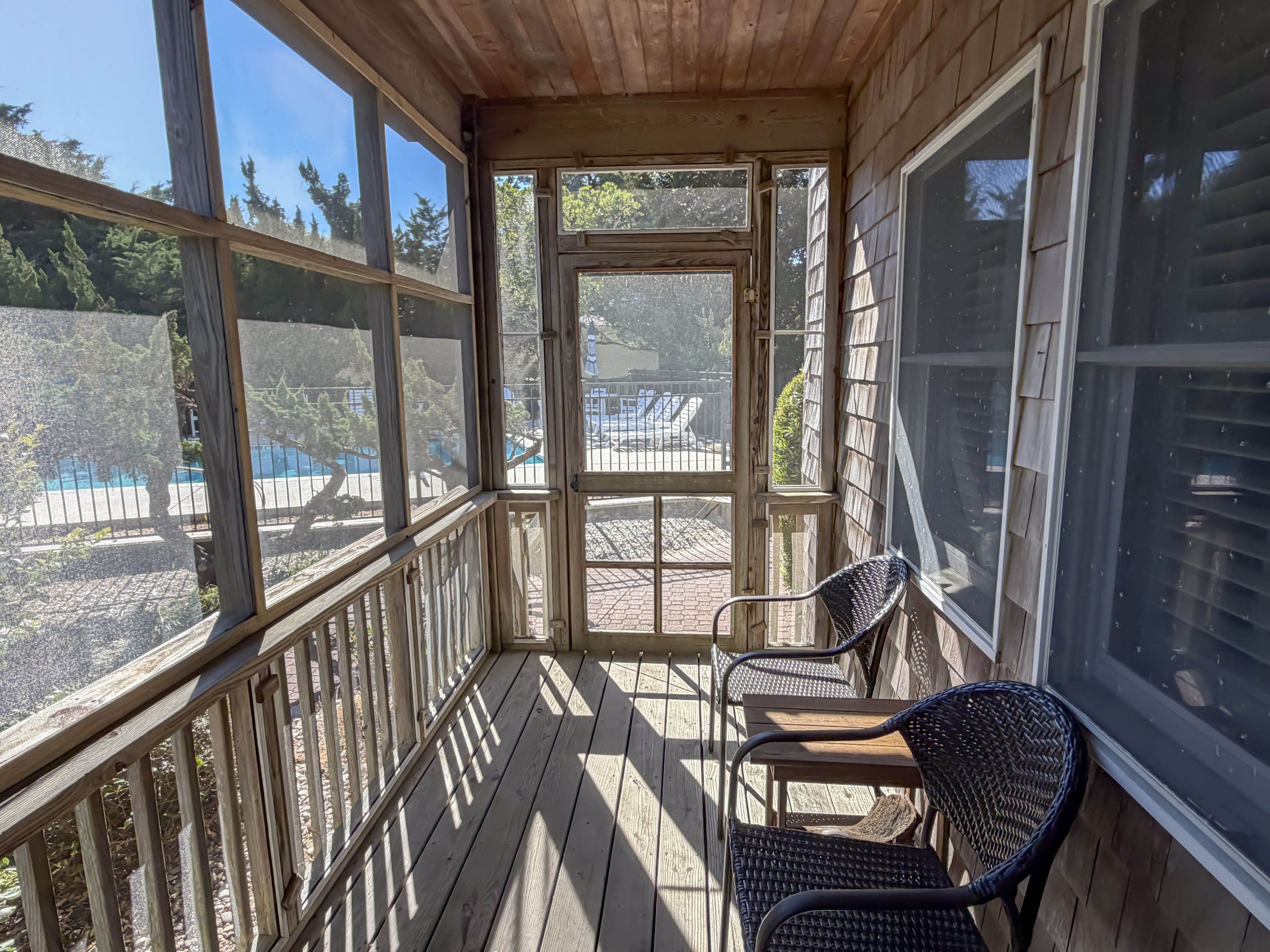 A wooden deck with a covered porch, two wicker chairs, and a view of a swimming pool and fenced yard.