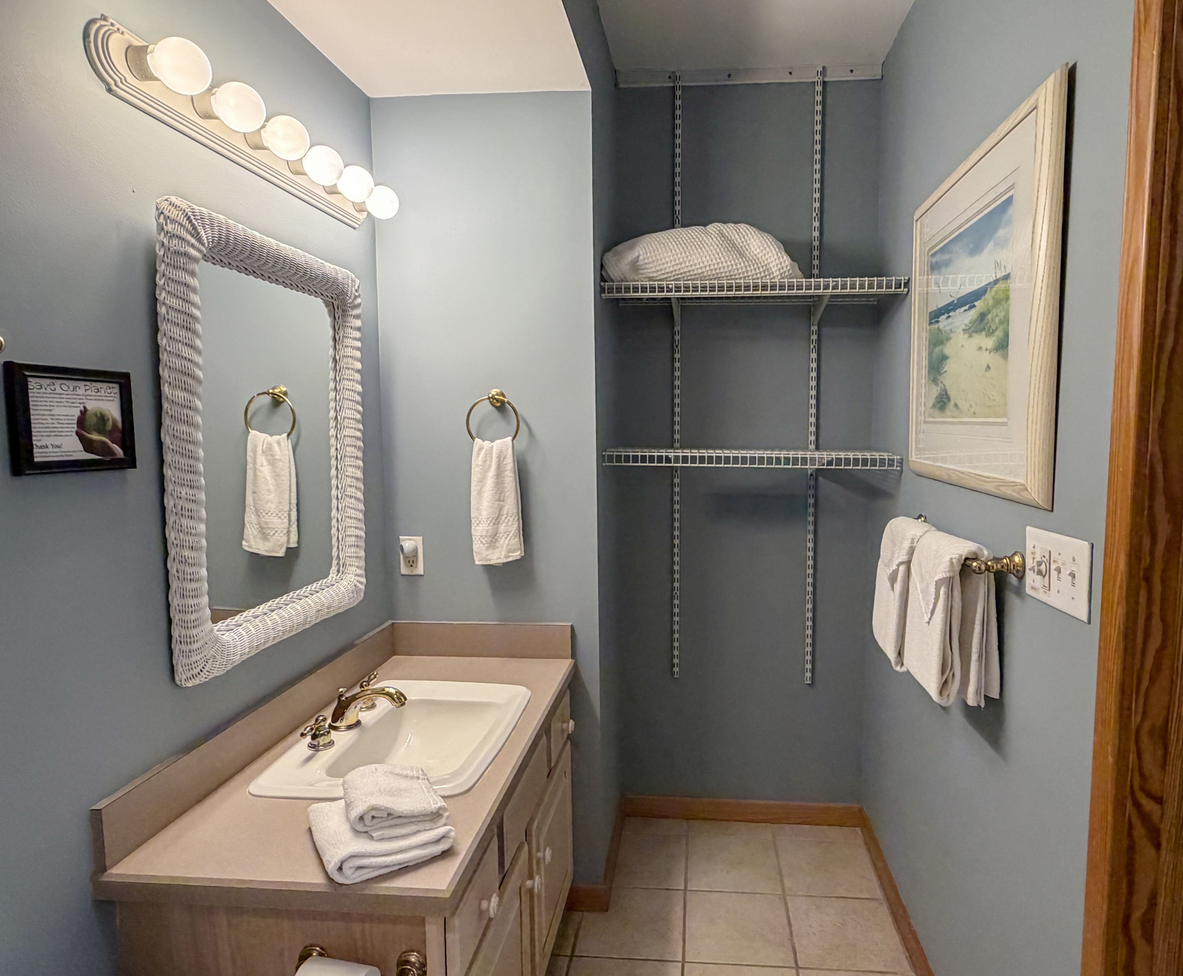 Un baño con tocador, un espejo grande, un estante en la pared y un cuadro enmarco.