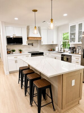 A modern kitchen with white cabinets, a large island with stools, stainless steel appliances, and pendant lighting.