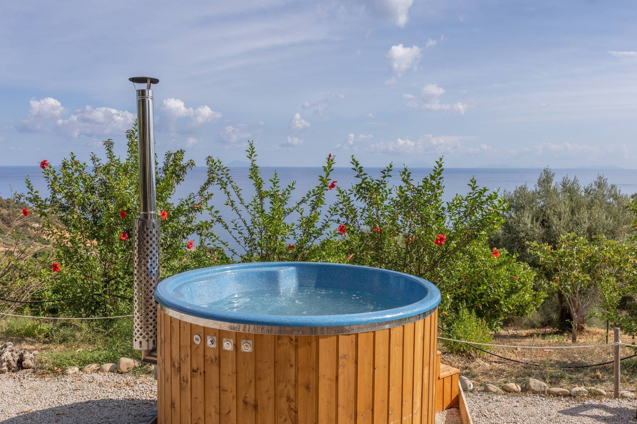 Ein Whirlpool mit Holzverkleidung und einem Metall-Heizelement, umgeben von viel Grün und mit Blick auf ein Gewässer.
