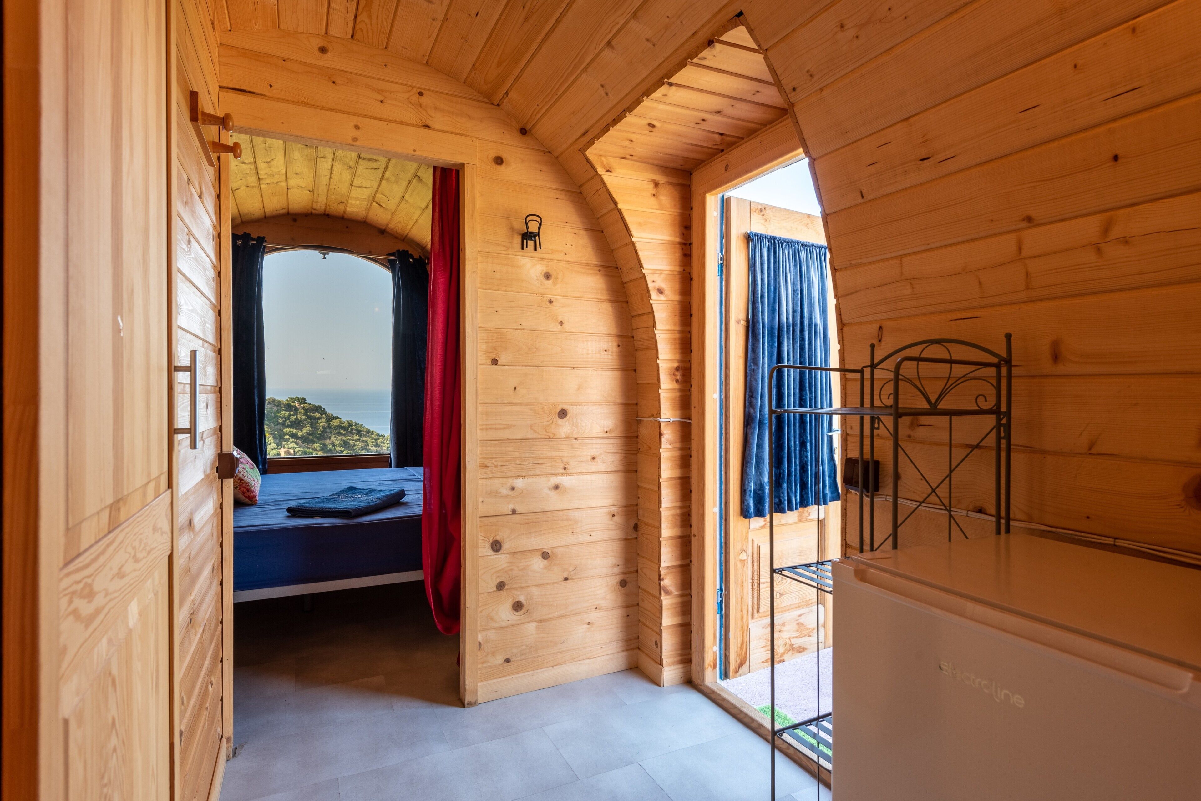 A wooden cabin interior with a bed, a view through an arched window, and a door leading to a balcony.