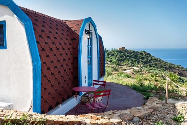 A building with a red and blue exterior, a small patio with a table and chairs, and a view of the sea and hills in the background.