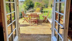Eine offene Holztür führt auf eine Terrasse mit Tisch und Stühlen, mit Blick auf eine grüne Landschaft mit Bäumen.