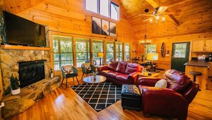A cozy cabin interior with a stone fireplace, leather sofas, a TV, and large windows offering a view of the outdoors. - New River Log Cabin | Waterfront • Sauna • Jet Tub (Pulaski County)